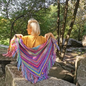 May include: A colorful crocheted shawl with fringe, featuring a rainbow of colors, including pink, purple, blue, green, yellow, and orange. The shawl is draped over a person's shoulders, with the fringe hanging down.