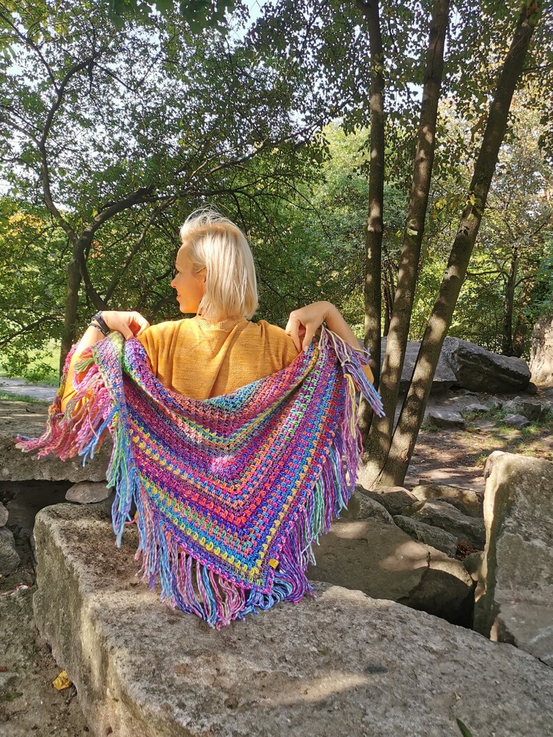 May include: A colorful crocheted shawl with fringe, featuring a rainbow of colors, including pink, purple, blue, green, yellow, and orange. The shawl is draped over a person's shoulders, with the fringe hanging down.