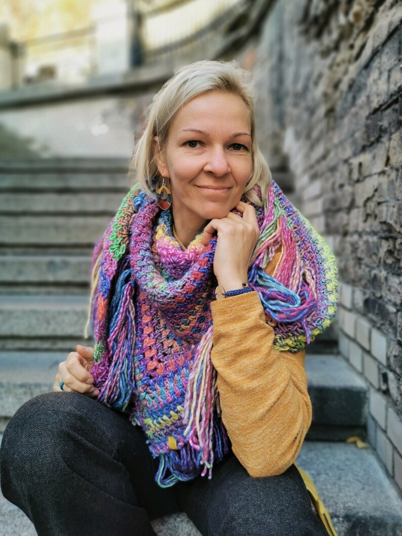 May include: A woman wearing a colorful crocheted shawl with fringe, a yellow sweater, and blue jeans. She is sitting on a set of stone steps.