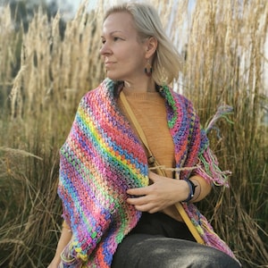 May include: A woman wearing a colorful crocheted shawl with a fringe, a mustard yellow sweater, and gray pants. She is sitting on a concrete surface with tall, dry grass in the background.