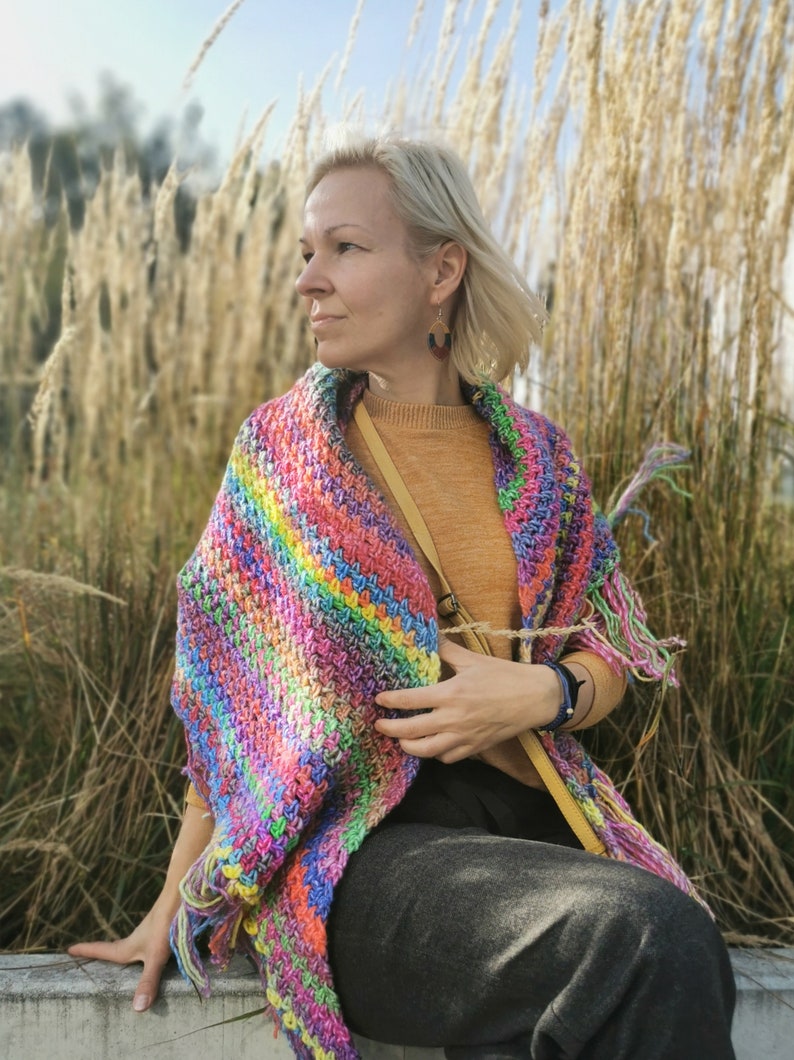May include: A woman wearing a colorful crocheted shawl with a fringe, a mustard yellow sweater, and gray pants. She is sitting on a concrete surface with tall, dry grass in the background.