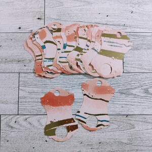 May include: A stack of pink, brown, and gold gift tags with a hole at the top. The tags have a watercolor-like design with stripes and abstract shapes.