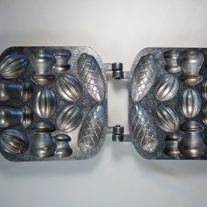 May include: A metal waffle iron with multiple molds for making different shapes of cookies or candies. The molds include nuts, acorns, and other shapes.