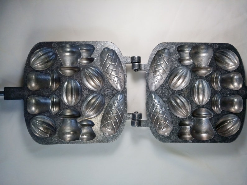 May include: A metal waffle iron with multiple molds for making different shapes of cookies or candies. The molds include nuts, acorns, and other shapes.
