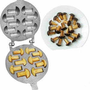 May include: A silver metal waffle iron with a handle. The waffle iron has 12 molds shaped like mushrooms. The waffle iron is open and shows a batch of golden brown mushroom-shaped waffles. A plate of chocolate-covered mushroom-shaped waffles is shown in the background.