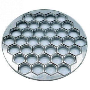 Pode incluir: Um molde redondo de metal prateado com 36 recortes circulares dispostos em um padrão hexagonal. O molde é usado para fazer bolinhos ou outras pequenas massas.