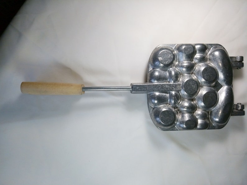 May include: A silver metal waffle iron with a wooden handle. The waffle iron has a pattern of round circles.