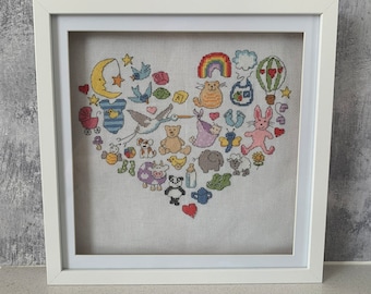 Puede incluir: Bordado de punto de cruz enmarcado en blanco de un corazón lleno de imágenes con temática de bebé, que incluyen una cigüeña, un biberon, un chupete, un sonajero, un osito de peluche, un globo aerostático, una luna, una estrella y un gato.