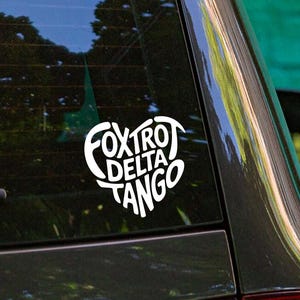 May include: A white heart-shaped vinyl decal applied to the rear window of a dark-colored vehicle. The decal features the text "FOXTROT DELTA TANGO" arranged in a stacked, stylized font within the heart outline, creating a clear graphic design.