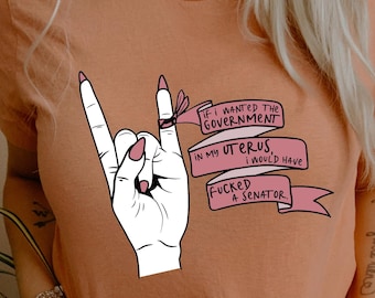 Könnte beinhalten: Ein pfirsichfarbenes T-Shirt mit einer Grafik einer weißen Hand, die eine Rock-and-Roll-Geste mit rosa Fingernägeln macht. Ein rosa Band wickelt sich um die Finger mit dem Text: IF I WANTED THE GOVERNMENT IN MY UTERUS, I WOULD HAVE FUCKED A SENATOR.