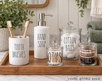May include: A white ceramic toothbrush holder labeled TOOTH BRUSHES, a white ceramic soap dispenser labeled HAND SOAP, and three clear glass jars with lids labeled Q TIPS, COTTON BALLS, and HAIR TIES, all arranged on a rectangular wooden tray.