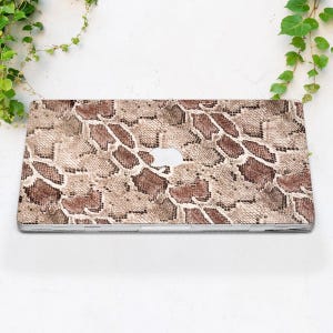 Puede incluir: Una funda para port&aacute;til con un dise&ntilde;o de piel de serpiente en tonos marr&oacute;n, beige y crema. La funda tiene un recorte para el logotipo de Apple y es de forma rectangular. El dise&ntilde;o imita la apariencia de la piel de reptil.
