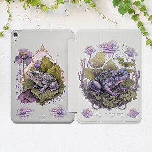 以下が含まれることがあります： グレーと白のiPadケースで、花柄とカエルのイラストが描かれています。ケースには画面を覆うフラップと、名前を入れるスペースがあります。カエルは紫と緑で、白い背景になっています。