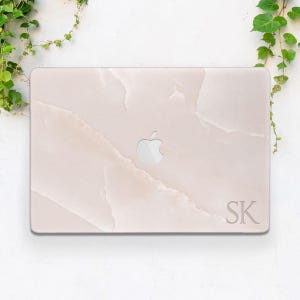 May include: A light pink laptop case with a marble design and the Apple logo. The case has the initials "SK" in the bottom right corner. Green vines are in the top left and right corners.