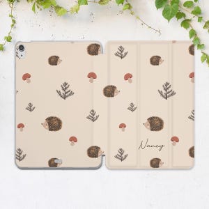 Può includere: Custodia beige per tablet con un motivo ripetuto di ricci marroni, funghi rossi e pini verde scuro. Il nome "Nancy" è scritto in corsivo sulla copertina. La custodia è aperta, rivelando lo schermo del tablet.