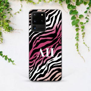 May include: A phone case with a zebra stripe pattern in black, white, and shades of pink. The initials "AH" are displayed in white block letters on the case. The phone case is shown against a white background with green vines.