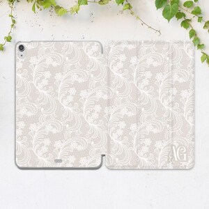 May include: A light beige tablet case featuring a white floral lace design. The case is open, displaying the tablet screen. The pattern includes delicate flowers and swirling vines. The initials "AG" are visible on the right side of the case.