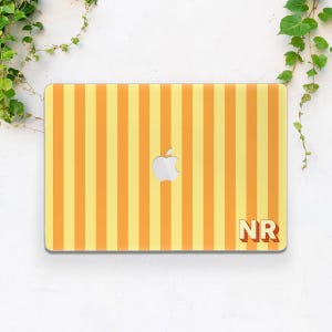 May include: A rectangular laptop skin featuring a vertical striped pattern in alternating shades of light yellow and orange. The center displays a silver Apple logo, and the bottom right corner features the bold, block-lettered text NR in orange.