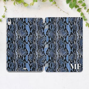 May include: A blue and black snakeskin-patterned tablet case, open to display the interior. The case has a hard shell design with a magnetic closure. The initials "MF" are displayed in white on the right side of the case.