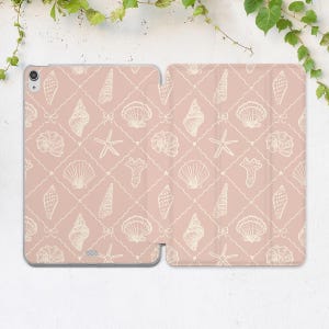 Può includere: Una custodia rosa per tablet con un motivo ripetuto di conchiglie bianche, stelle marine e fiocchi. Il design è ambientato su uno sfondo a griglia, creando un tema nautico. La custodia è aperta, mostrando il design interno ed esterno.