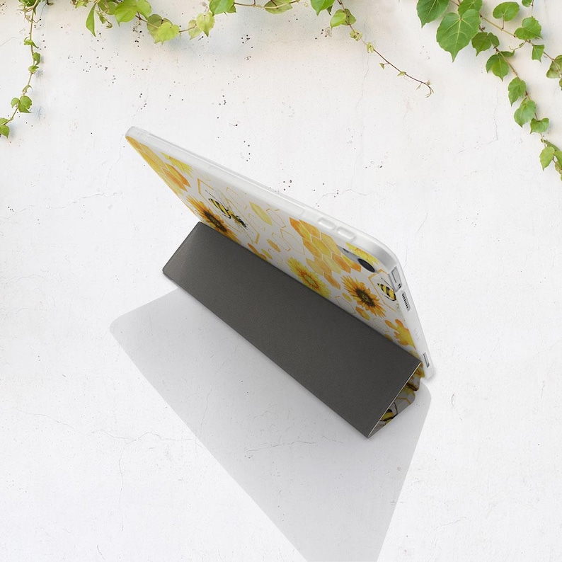 K&ouml;nnte beinhalten: Eine wei&szlig;e Tablet-H&uuml;lle mit gelbem Sonnenblumenmuster wird von einem grauen St&auml;nder gehalten. Die H&uuml;lle hat einen transparenten Hintergrund mit gelben Sonnenblumen und schwarzen Bienen. Der St&auml;nder hat eine dunkelgraue, abgewinkelte Form.