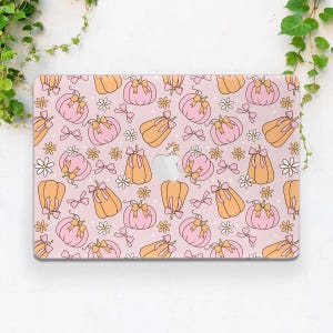 May include: A pink laptop case featuring a repeating pattern of pumpkins in shades of pink and orange, adorned with bows and white daisy-like flowers. The case has a cutout for the Apple logo.