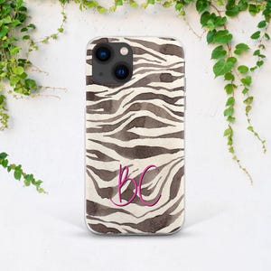 May include: A clear phone case with a zebra print design in shades of brown and white. The case features the pink initials "BC" in a cursive font. The phone case has a camera cutout and is surrounded by green vines.