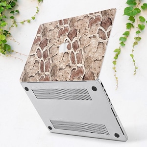 Puede incluir: Un ordenador port&aacute;til con una funda con estampado de piel de serpiente en tonos marr&oacute;n y beige. El port&aacute;til est&aacute; abierto, revelando la parte inferior plateada con ranuras de ventilaci&oacute;n. Hojas verdes en el fondo.