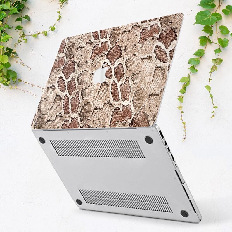 Puede incluir: Un ordenador port&aacute;til con una funda con estampado de piel de serpiente en tonos marr&oacute;n y beige. El port&aacute;til est&aacute; abierto, revelando la parte inferior plateada con ranuras de ventilaci&oacute;n. Hojas verdes en el fondo.