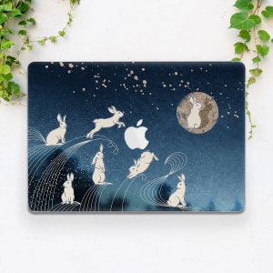 以下が含まれることがあります： 星が降る夜空のデザインが施された青いノートパソコンのスキンの上に、白いウサギと月が描かれています。スキンには銀色のアップルのロゴがあります。