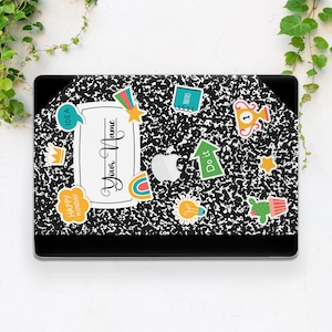 May include: A black and white laptop skin with a notebook paper design. The skin features colorful stickers with various designs, including a rainbow, a lightbulb, a crown, a star, a trophy, a cactus, and a speech bubble that says "Happy Monday".