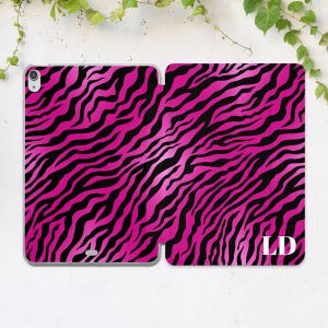 May include: A hot pink and black zebra print tablet case, open to display the interior. The case features a bold, eye-catching pattern with the initials "LD" in white on the right side. The case is designed to protect a tablet.