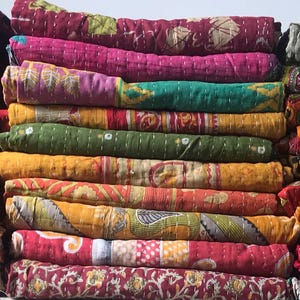 Puede incluir: Montones de colchas coloridas y estampadas en varios tamaños y diseños. Las colchas presentan una mezcla de patrones florales, geométricos y abstractos en tonos de rojo, rosa, naranja, verde y azul. Las colchas están dobladas y apiladas, mostrando sus colores vibrantes.
