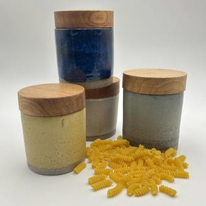 May include: Four ceramic canisters with wooden lids in shades of blue, beige, yellow, and green. A pile of uncooked fusilli pasta is in front of the canisters. The canisters are cylindrical and have a rustic, handmade appearance.