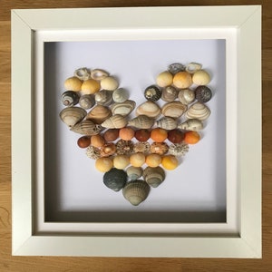 May include: A white frame with a white mount surrounding a heart shape made of seashells. The seashells are arranged in a gradient of colours, from light yellow to dark brown.