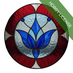 May include: Circular stained glass window with a floral design. The window features a central blue flower, surrounded by light blue and clear glass. A red border and accents complete the design. The words "HOBBY LICENSE" are printed on a green diagonal banner.