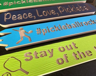 May include: Four wooden signs with different messages and designs. The top sign is navy blue with white text that reads "Peace, Love, Pickleball". The second sign is light blue with a white silhouette of a person playing pickleball and the text "#pickleballrocks". The third sign is green with a white silhouette of a pickleball paddle and the text "Stay out of the k".