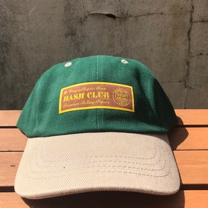 May include: A green and beige baseball cap with a brown patch that says "Kingsize Organic Hemp Jash Club Premium Rolling Papers" and a smaller brown patch that says "Jash Club".