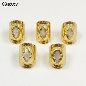 May include: Five gold-toned rings arranged in a group, each featuring a smooth, polished metal band with a central oval cutout. Inside each cutout is a white, iridescent mother-of-pearl inlay shaped like a four-pointed cross. The WKT logo is visible.