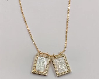 May include: Gold-toned necklace featuring two rectangular pendants. Each pendant has a white mother-of-pearl inlay with a religious image, framed by a row of small, clear crystals. The necklace has a delicate chain and the logo "WKT" in the upper left corner.