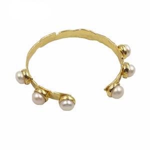 May include: A gold-coloured cuff bracelet with a hammered texture. The open-ended bracelet features six white pearls set in gold-coloured bezels. The bracelet has a unique, organic design, suitable for everyday wear or special occasions.
