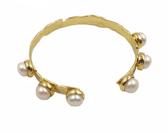 May include: A gold-colored cuff bracelet with a hammered texture. The open-ended bracelet features six white pearls set in gold-colored bezels. The bracelet has a unique, organic design, suitable for everyday wear or special occasions.