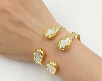 Può includere: Un bracciale rigido color oro con quattro castoni ovali e inserti in conchiglia bianca. La superficie metallica presenta una finitura martellata e testurizzata. Il design è composto da un bracciale aperto con due elementi decorativi per lato.