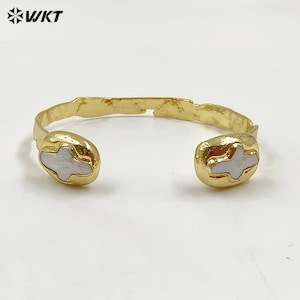 Könnte beinhalten: Ein goldenes Armreif-Armband mit zwei weißen, kreuzförmigen Akzenten. Das Armband ist offen und hat eine gehämmerte Textur.