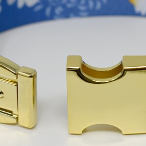 May include: Close-up of a gold metal dog collar buckle. The collar is blue with a white floral pattern.