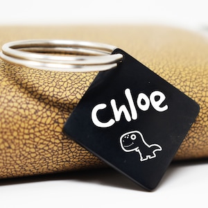 May include: Black square keychain with a white dinosaur and the name Chloe written in white.
