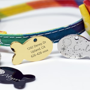 May include: Three gold and silver fish-shaped pet ID tags with engraved text. The gold tag has an address and phone number. The silver tag has a moon and stars design and the name Carla. The black tag has a yarn ball design and the name Alyssa.