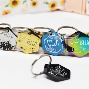 May include: Five different hexagonal dog tags with the name "Bella" engraved on each. The tags are attached to silver key rings and feature various designs, including mountains, stars, flowers, a cat, and a cloud.