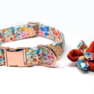 May include: A white dog collar with a floral pattern and a rose gold buckle. A matching rose gold D-ring is attached to the collar. A red and white floral bow is included.