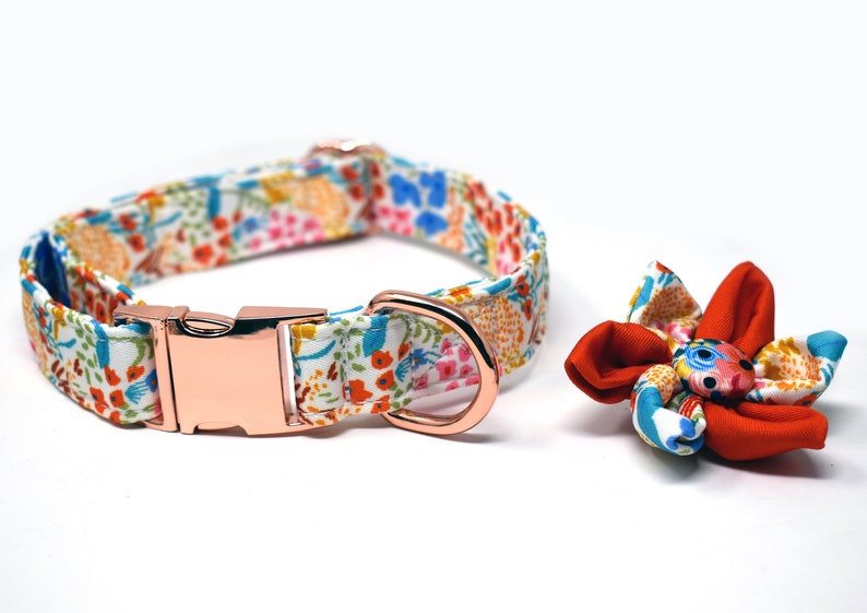May include: A white dog collar with a floral pattern and a rose gold buckle. A matching rose gold D-ring is attached to the collar. A red and white floral bow is included.